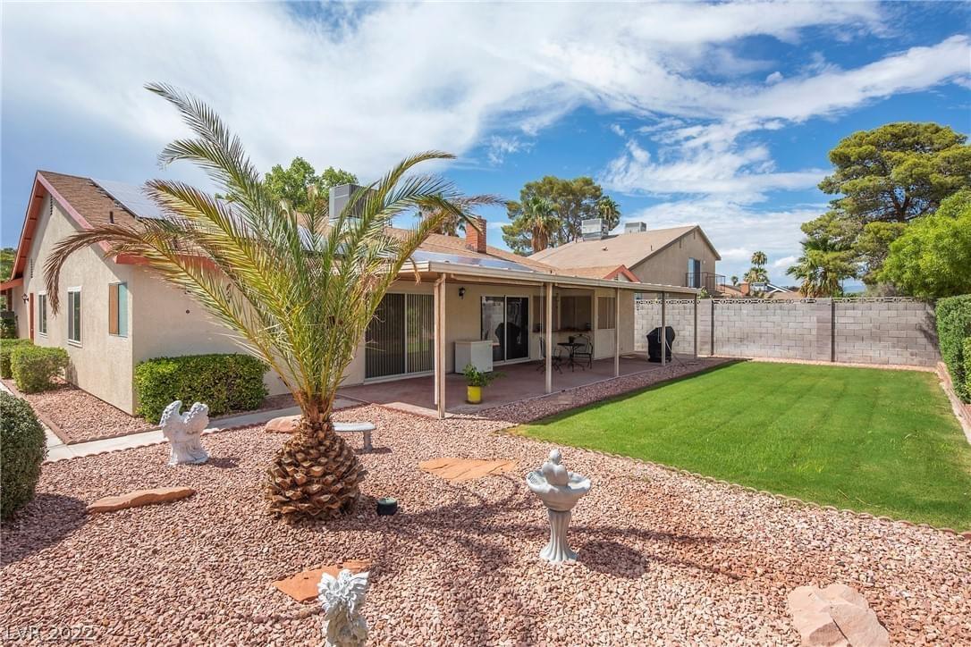 A single-story, light-colored stucco home with covered porch, plot of grass, gravel landscaping with palm tree, and solar panels